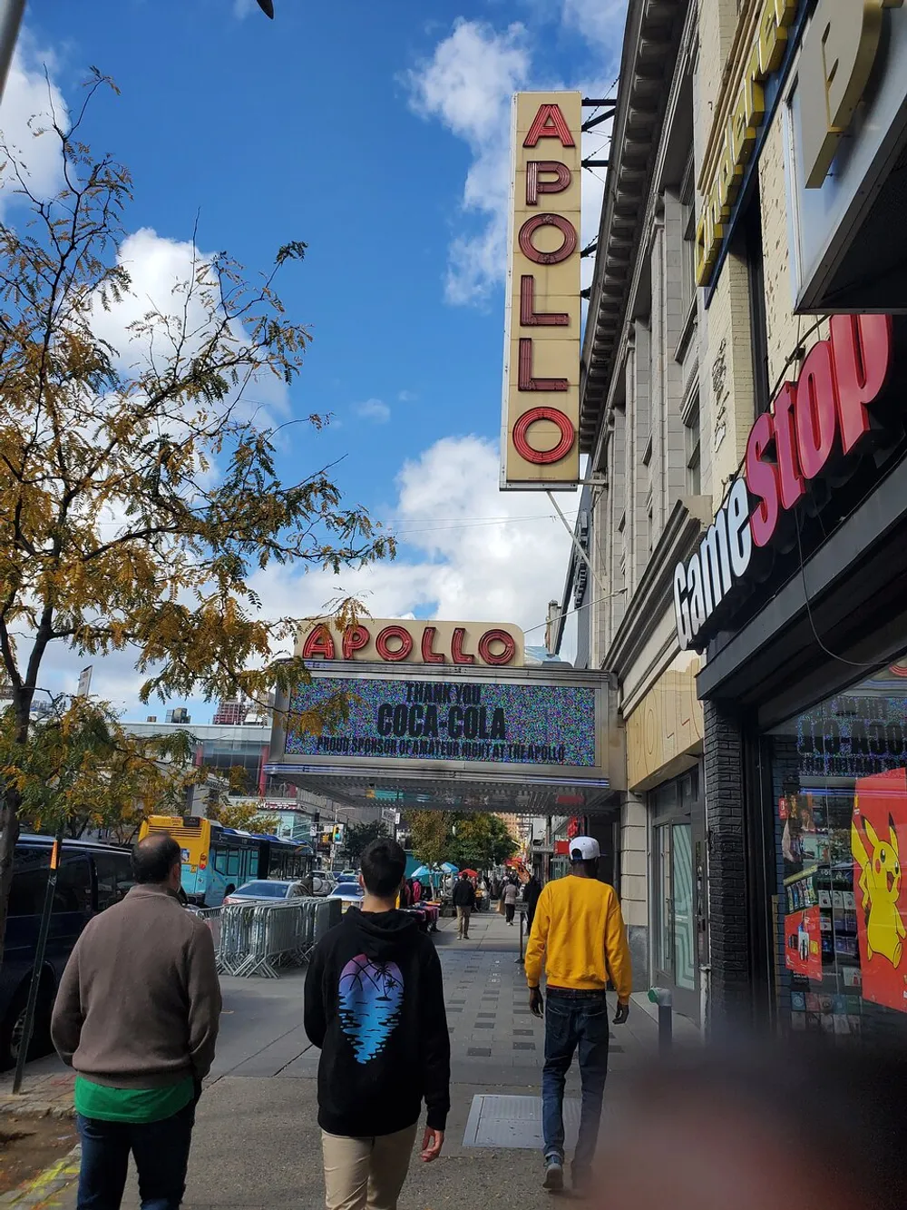 The image shows pedestrians walking past the iconic Apollo Theater with its prominent signage under a partly cloudy sky
