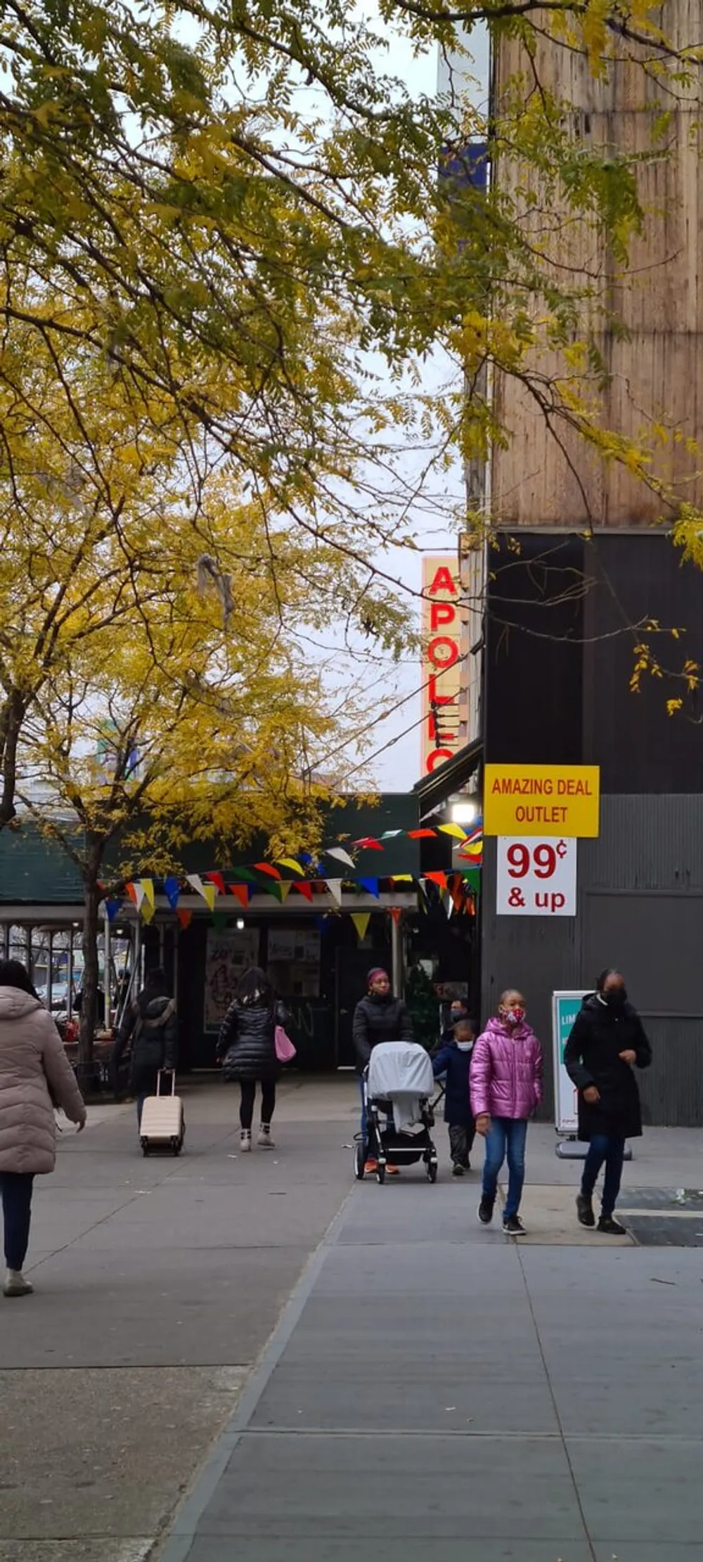This image captures a bustling city sidewalk scene with pedestrians autumnal trees and a store with a sign advertising an AMAZING DEAL 99  up under a marquee that reads APOLLO