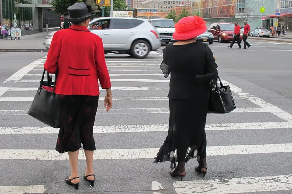 Two elegantly dressed individuals one in a red outfit and the other in black are standing at a pedestrian crossing in an urban setting