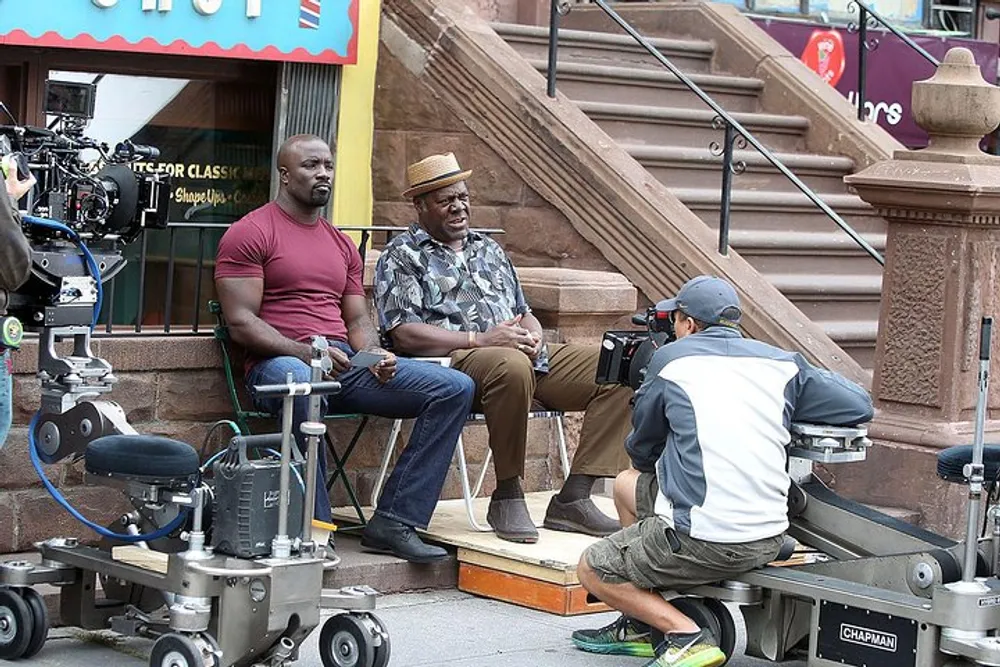 Two actors are seated on a stoop during a film or television shoot while a camera operator captures the scene