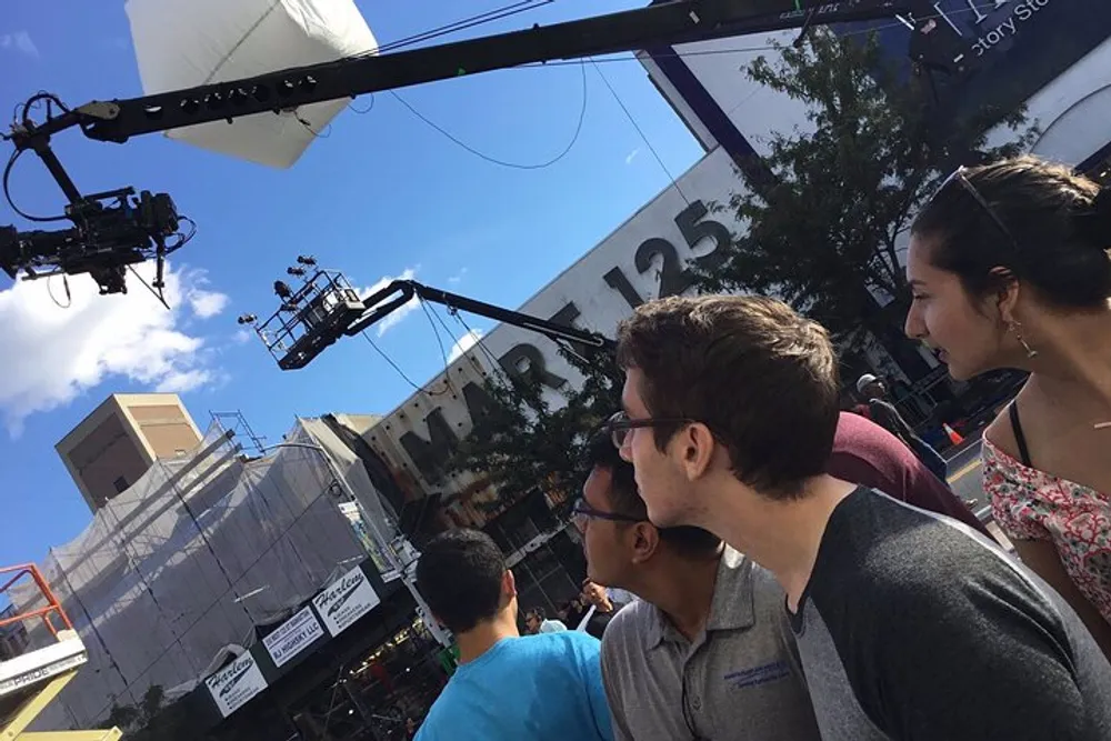 The image shows a group of people looking upwards at a film or television production setup on a sunny day with a crane-mounted camera and scaffolding in view