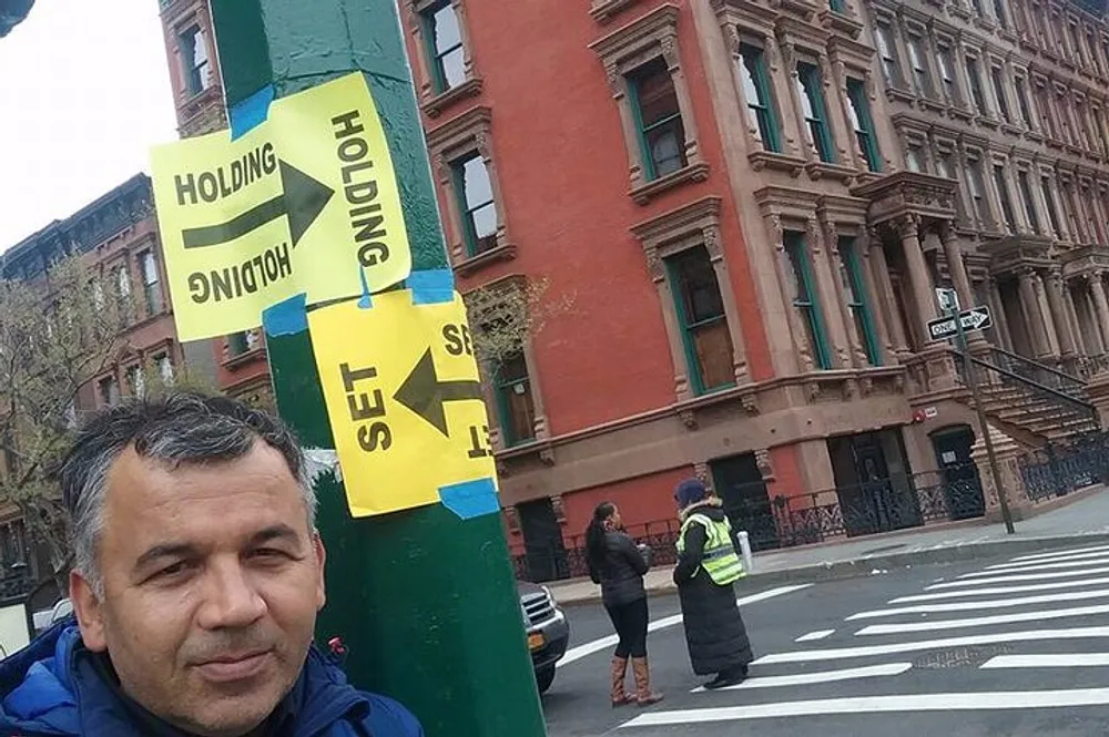A person is taking a selfie on an urban street corner with HOLDING SET signs attached to a post with other individuals and a crosswalk in the background