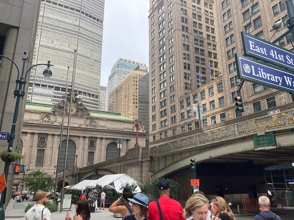 The image depicts a bustling city street corner with pedestrians diverse architecture including a historic building modern skyscrapers vehicles and street signs indicating East 41st Street and Library Way