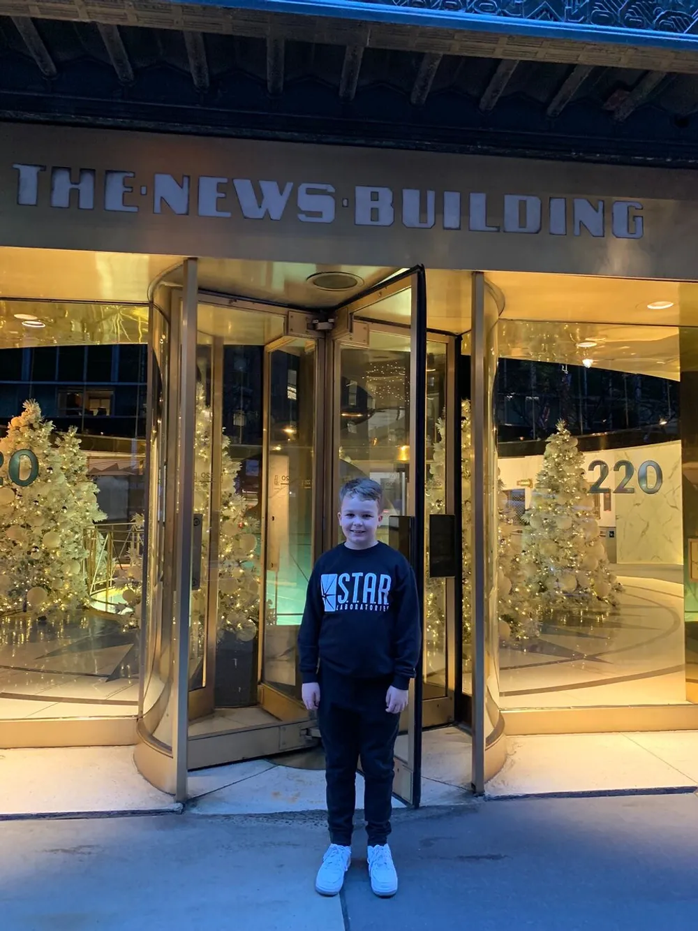 A young person is standing in front of the entrance to The News Building decorated with festive trees under an illuminated sign