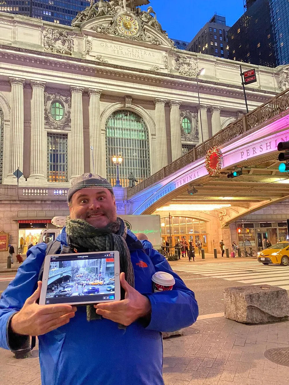 A person stands smiling in front of Grand Central Terminal at night holding a tablet that displays an image of a busy city street