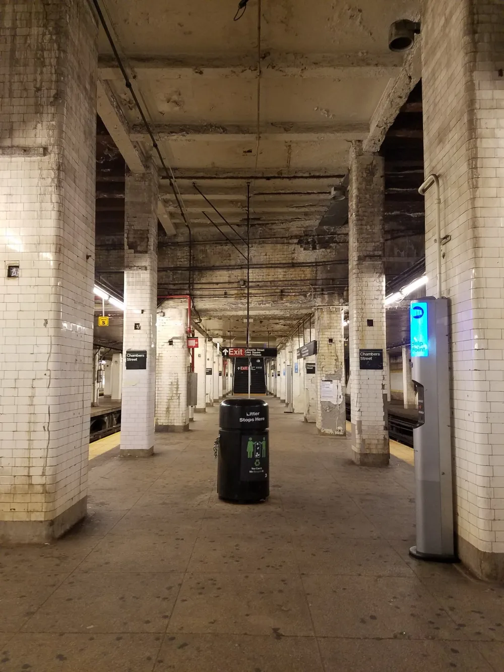 The image shows a seemingly empty aged subway station platform with white tiled walls an overhead structure exposing beams and pipes a trash bin prominently placed in the center and signage including a lit Exit sign