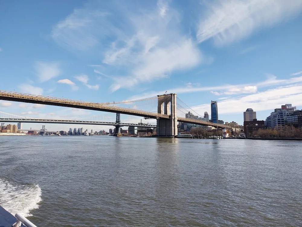 The image features a panoramic view of the Brooklyn Bridge stretching across the East River with a clear blue sky above and part of a watercrafts railing visible in the foreground