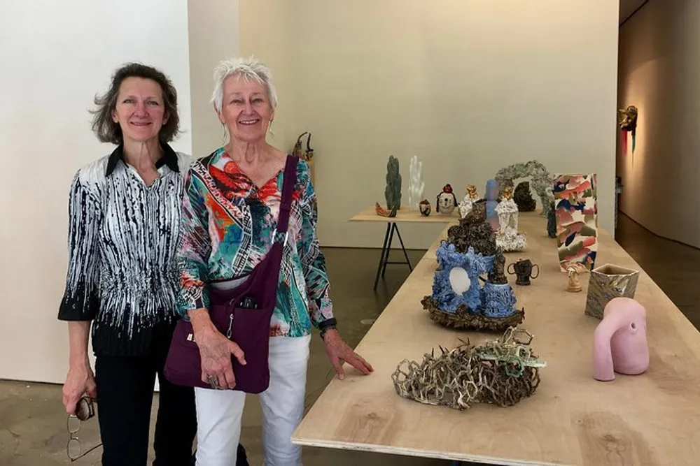 Two women are smiling for a photo while standing next to a table displaying an eclectic array of art pieces in a bright gallery setting