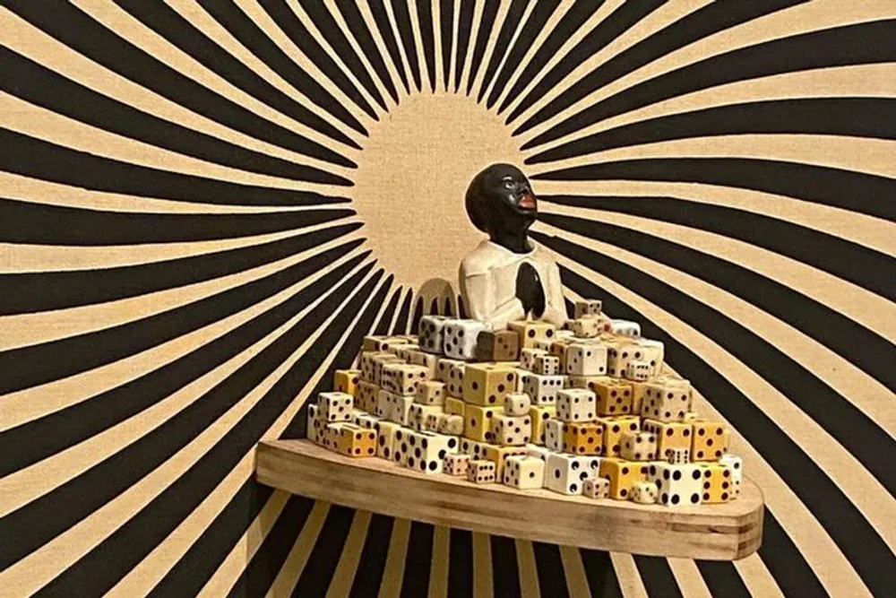 A sculpture of a seated figure surrounded by dice is set against a backdrop with a radial black and white design