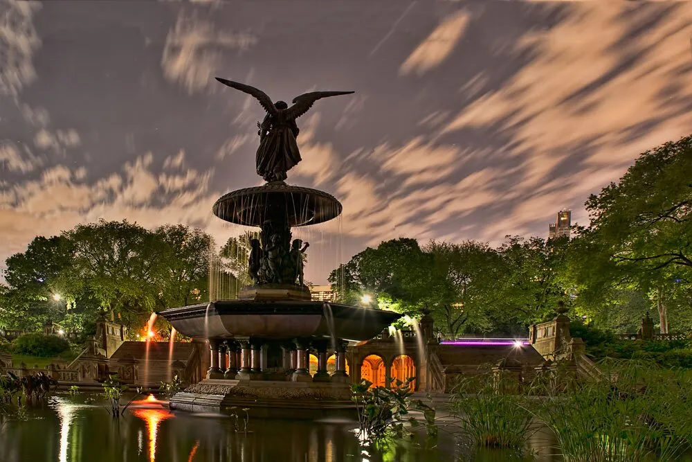 A long-exposure photograph captures the dynamic movement of clouds over the lit-up Bethesda Fountain in Central Park at night