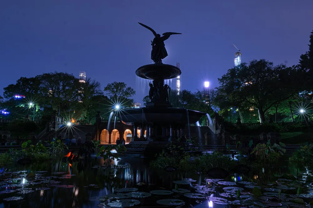 The image shows a beautifully illuminated fountain at night set amidst a serene pond with lily pads with glowing city lights and buildings in the background creating a juxtaposition of natural tranquility and urban energy