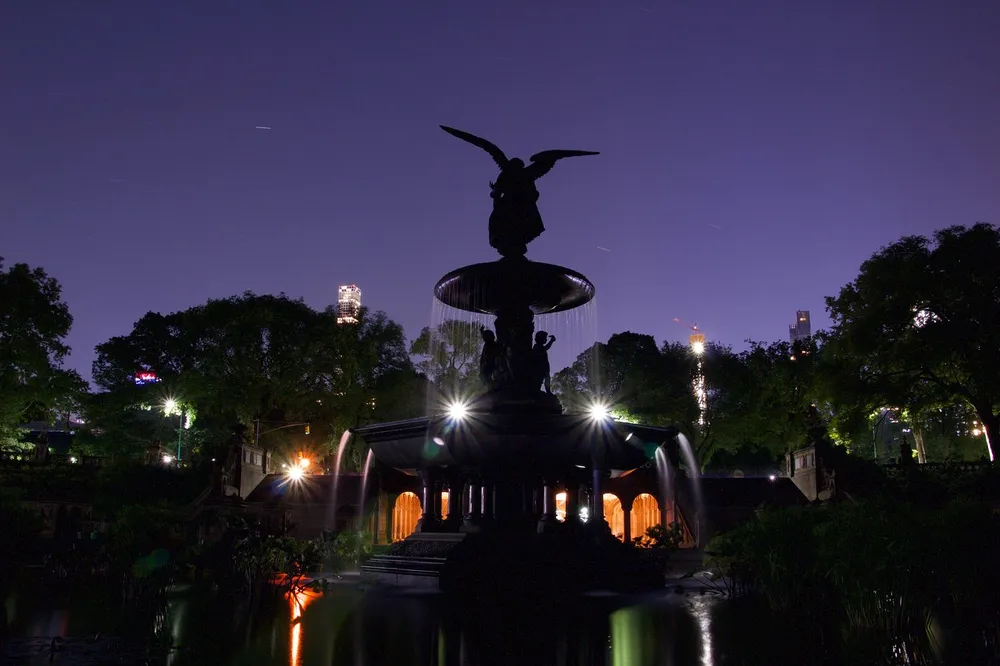 The image captures a silhouette of the Bethesda Fountain at twilight with star trails in the night sky and the illuminated cityscape in the background