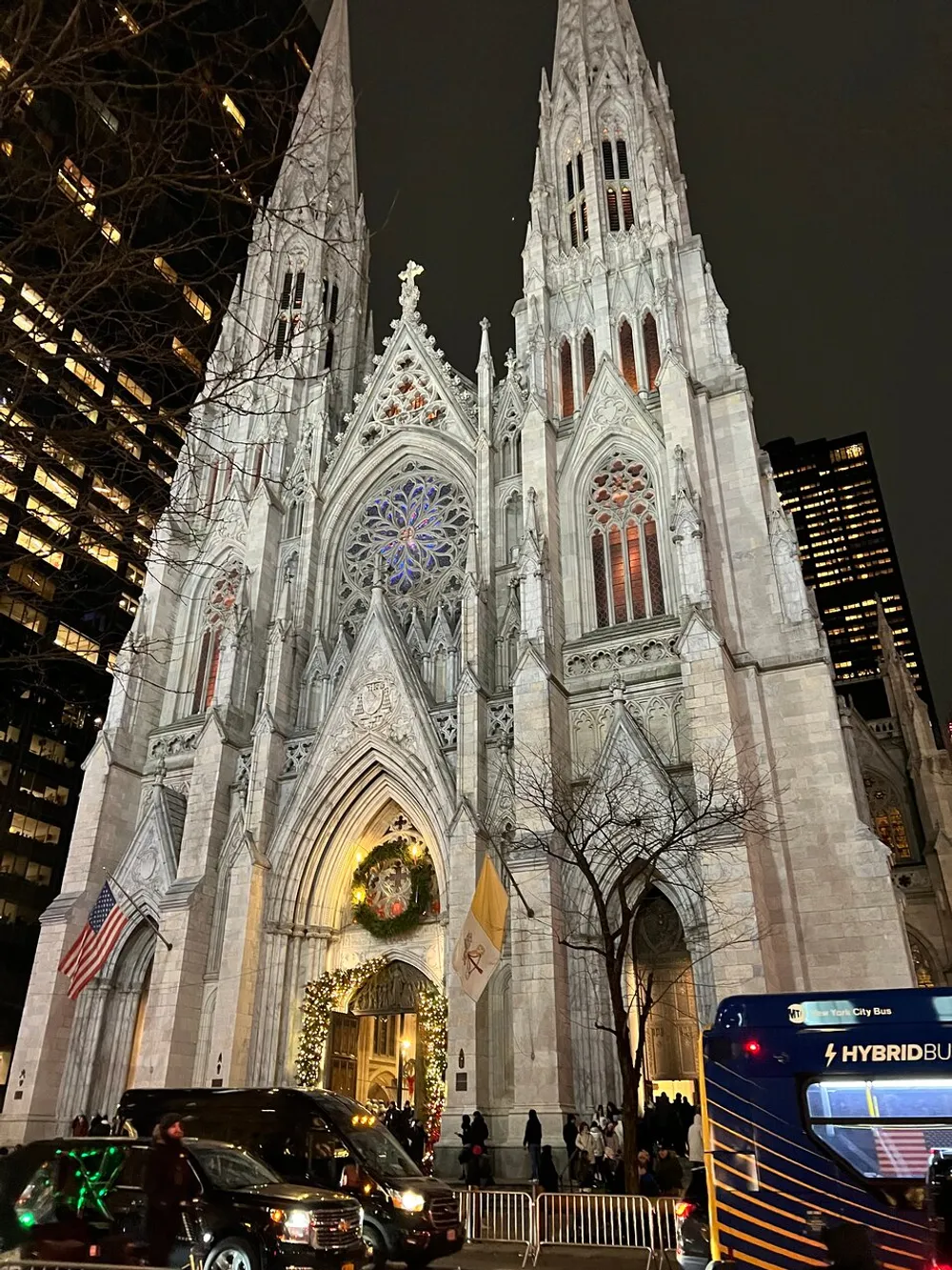 The image captures a grand Gothic cathedral illuminated at night adorned with a large wreath and festive decorations as traffic and pedestrians navigate the bustling street alongside it