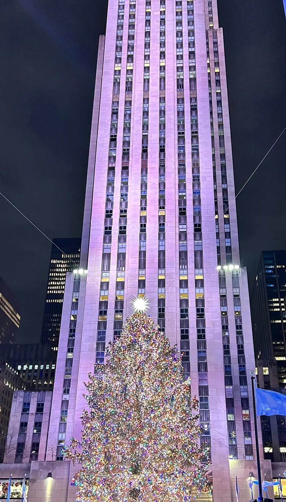 A large festively lit Christmas tree stands before a towering skyscraper illuminating the plaza at night