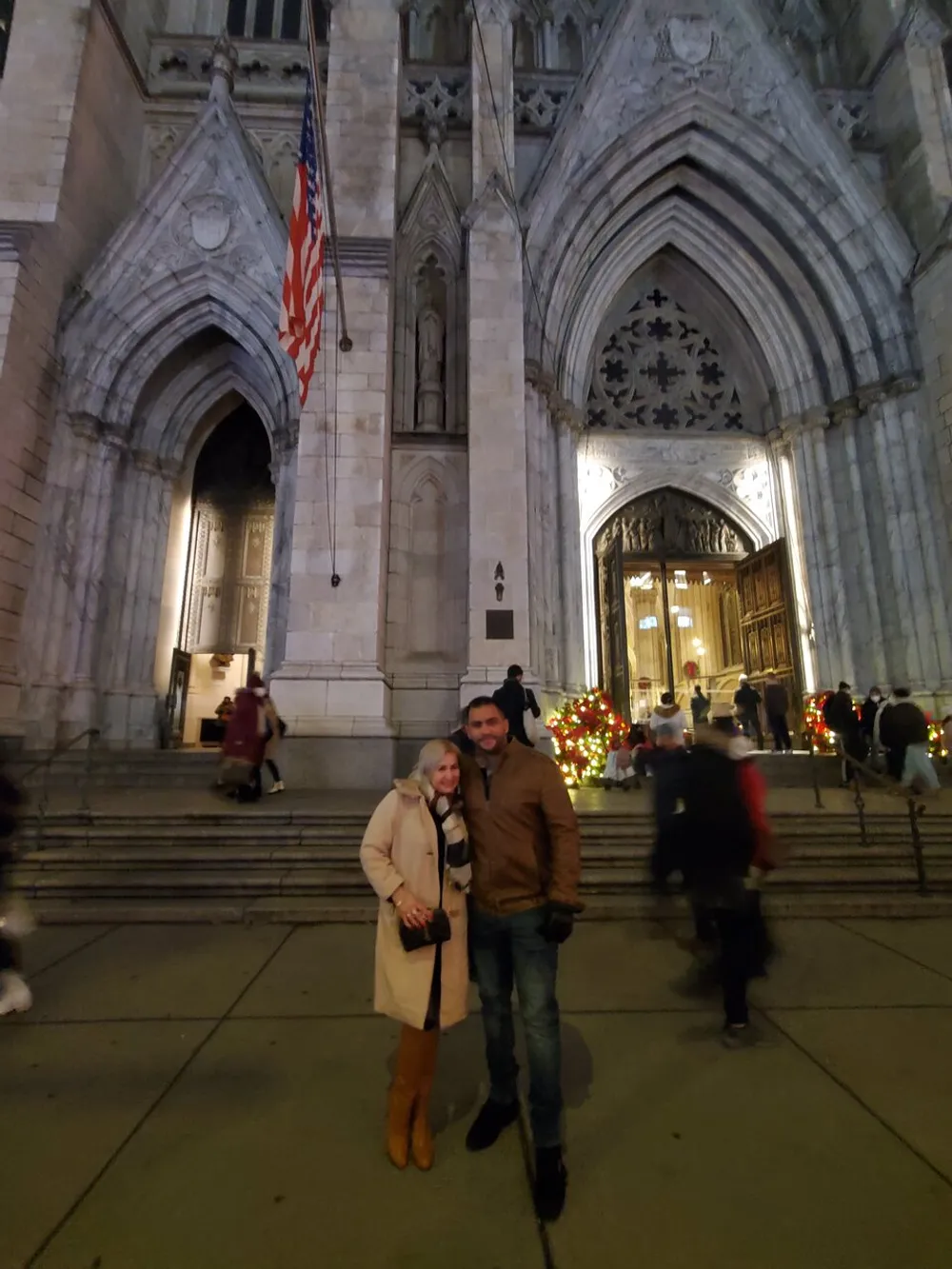 A couple is posing for a photo in front of an ornately designed cathedral entrance adorned with an American flag and holiday decorations
