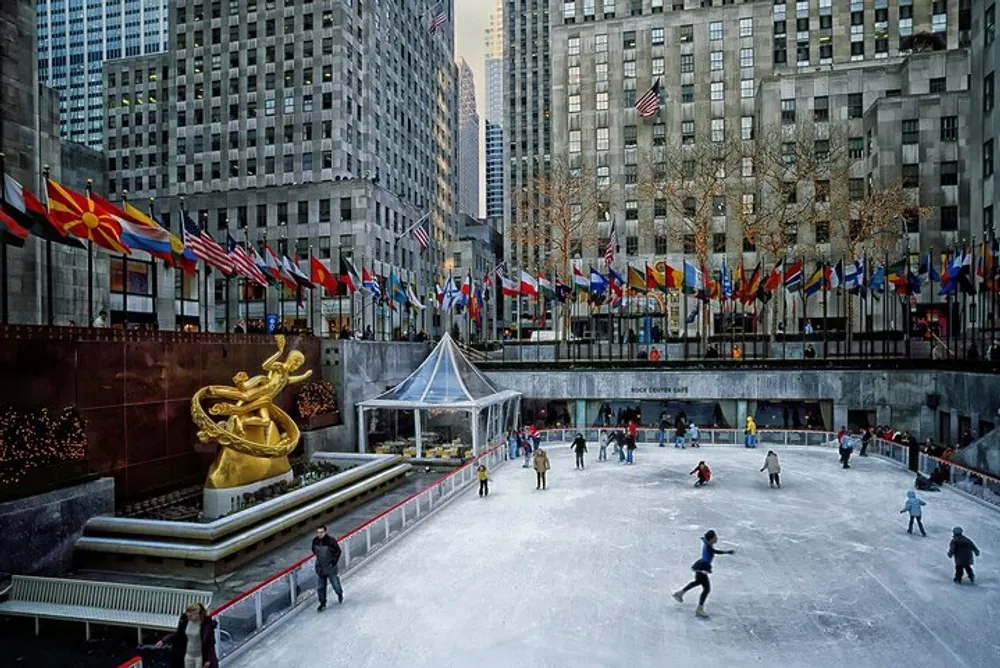 The image showcases an outdoor ice-skating rink surrounded by tall buildings with a golden statue to the left a row of international flags and people skating or walking around the rink
