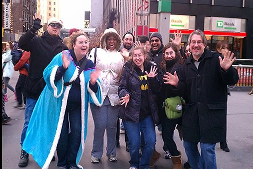 A group of cheerful people posing for a photo on a busy city sidewalk with some raising their hands or making gestures to the camera