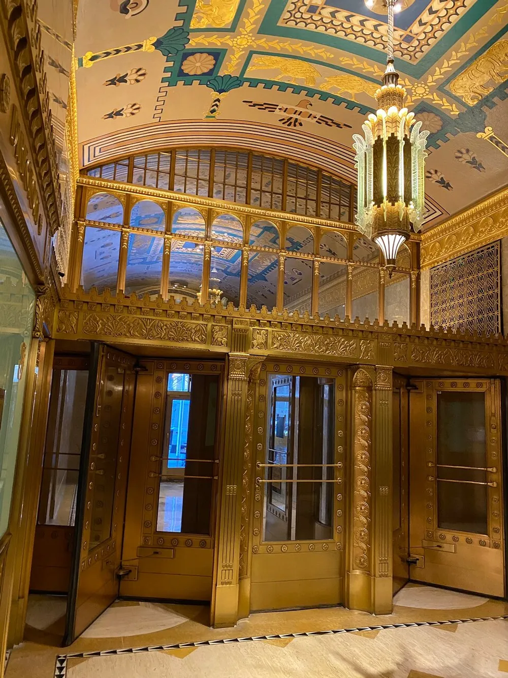 The image shows an ornate Art Deco interior with golden elevator doors decorative panels and a detailed ceiling with a hanging chandelier reflecting a luxurious architectural style