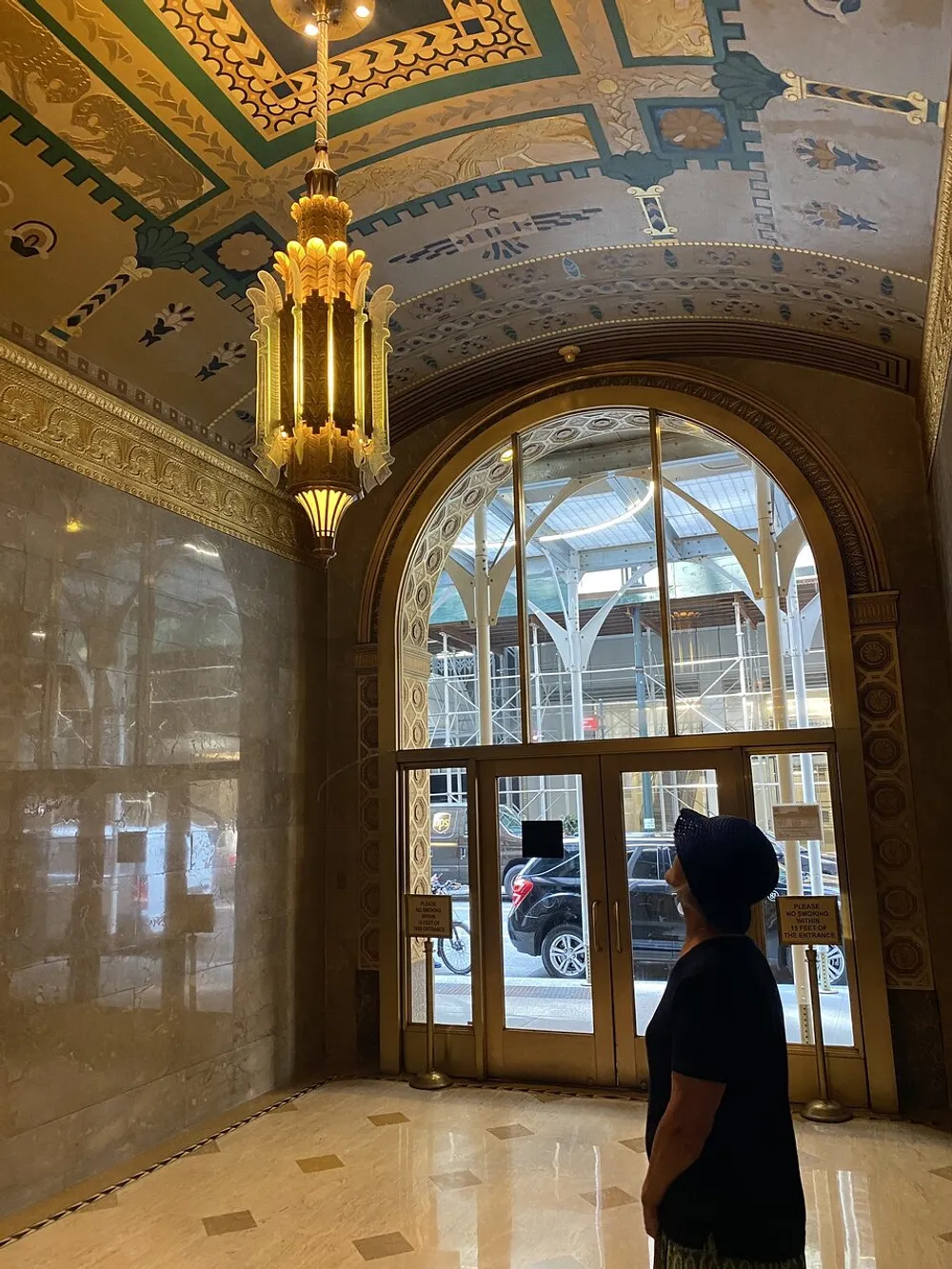 A person is standing inside a grand building with ornate ceilings and a chandelier looking out through arched glass doors