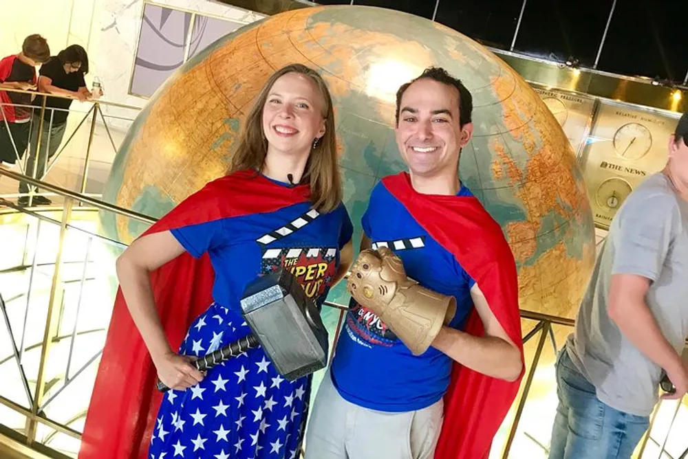 Two individuals are smiling for the camera with one dressed in a star-spangled outfit and the other in a superhero-themed shirt both wearing red capes and posing in front of a large globe structure