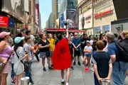 A performer wearing a red cloak is engaging with a crowd of onlookers in a bustling Times Square.
