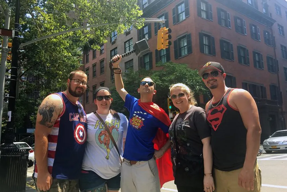 A group of five people are posing on a sunny street corner four of whom are wearing superhero-themed clothing including Captain America The Flash Superman and a Superman logo tank top while striking a friendly and casual pose