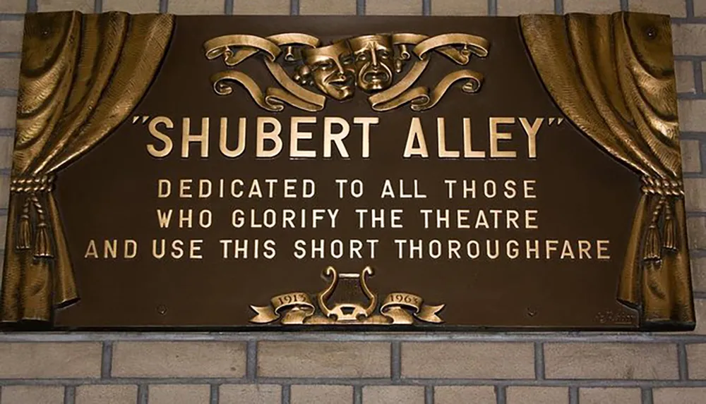 The image shows a commemorative plaque inscribed with SHUBERT ALLEY and a dedication adorned with theatrical masks and a curtain motif symbolizing its significance to the theater community