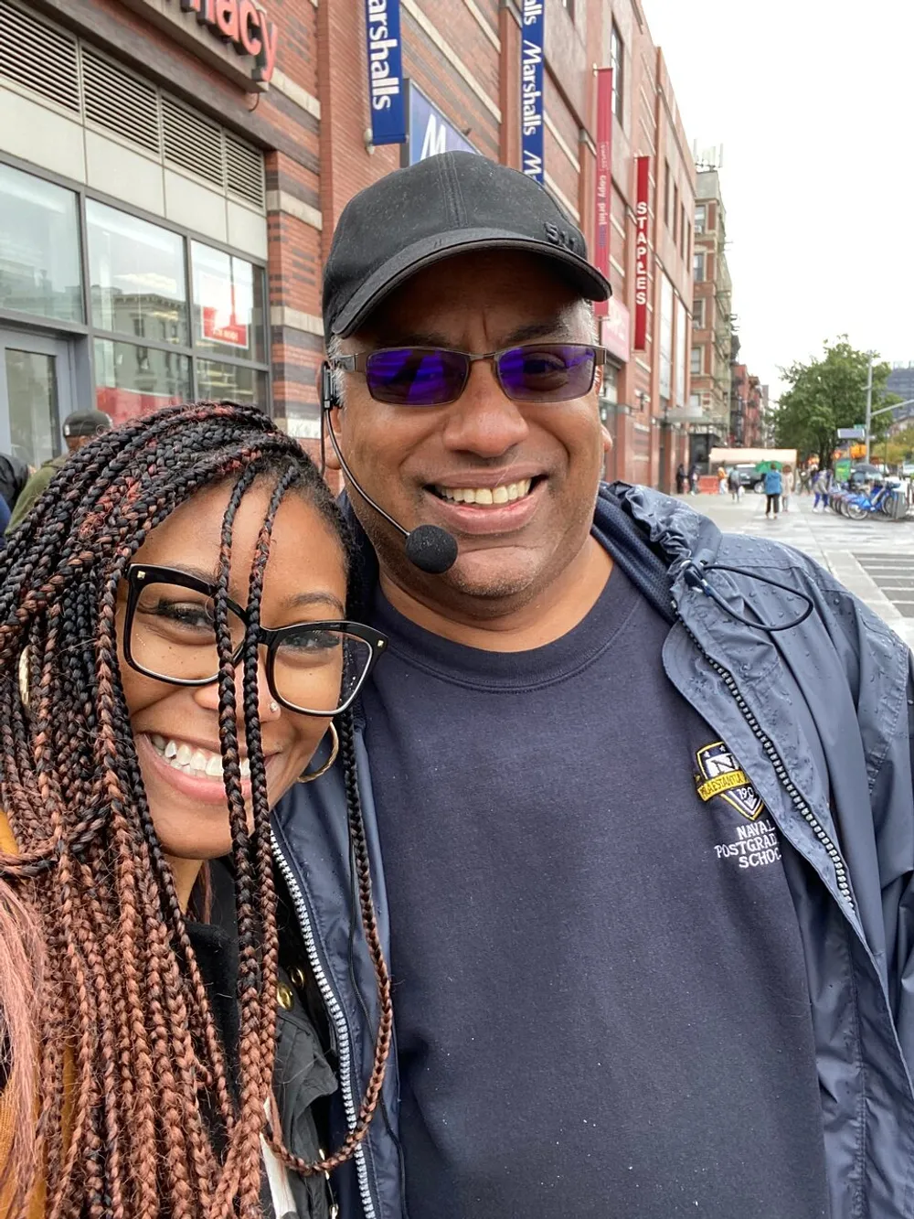 A woman and a man with a headset are smiling for a selfie on a city street with commercial buildings in the background