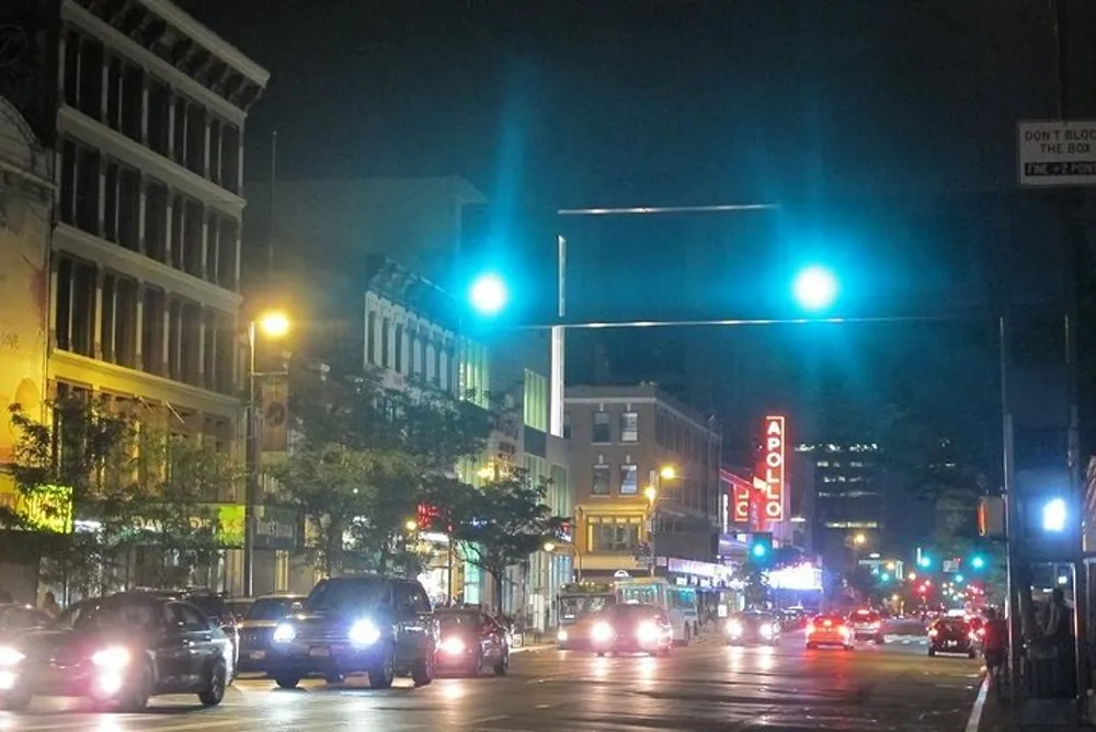 The image shows a bustling urban street at night highlighted by the illuminated signage of the Apollo Theater with cars on the road and a dynamic cityscape backdrop