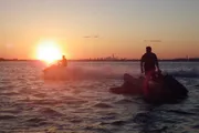 Two individuals are riding jet skis on a body of water at sunset with a city skyline visible in the background.