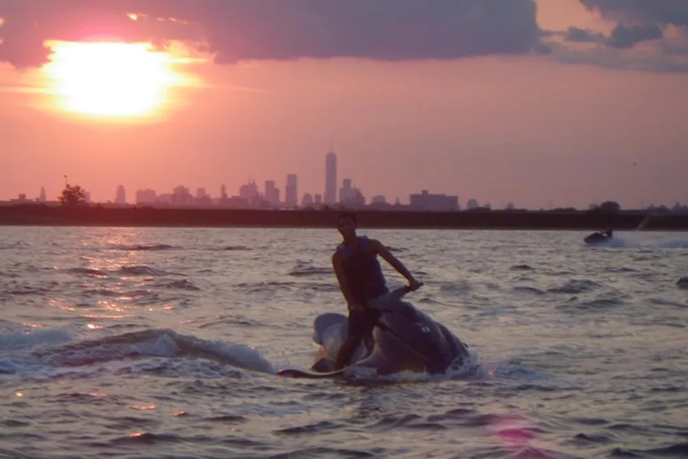 A person is riding a jet ski on the water at sunset with a city skyline in the background