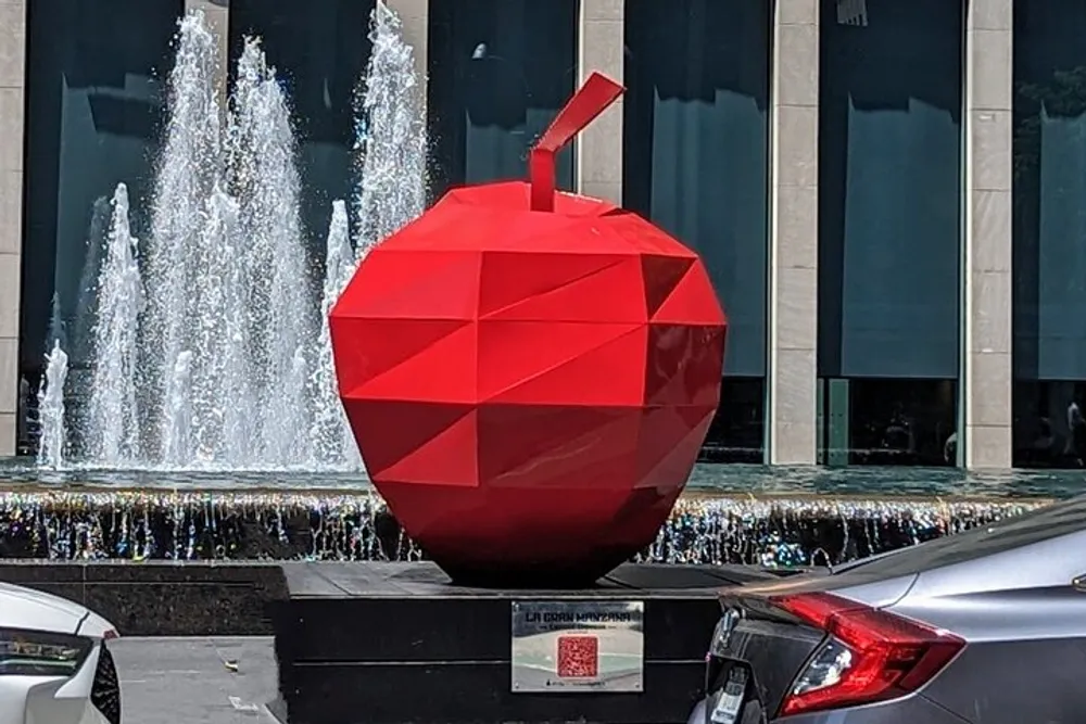 The image shows a large red geometric apple sculpture displayed in an urban setting with a fountain in the background