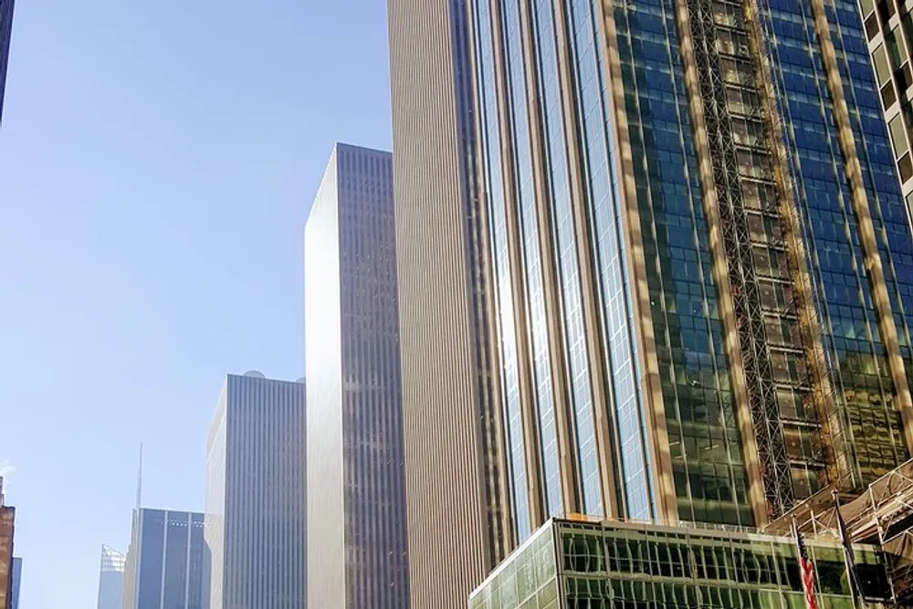 The image shows a group of modern skyscrapers rising against a clear blue sky reflecting sunlight off their glass facades