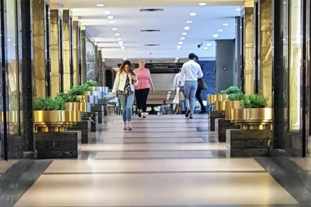 The image shows a modern interior hallway with people walking featuring reflective floors decorative plants in golden pots and dark marble walls