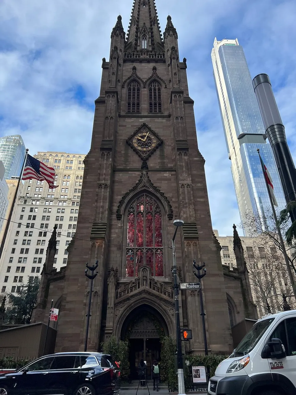 The image shows a historic stone church with a large gothic arch and stained glass window located on the corner of Broadway juxtaposed against the backdrop of modern skyscrapers and an American flag fluttering in the city
