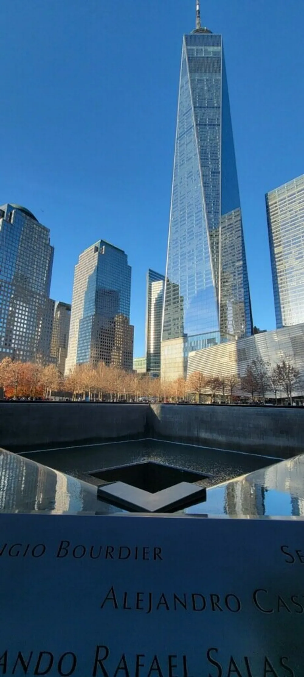 The image displays the One World Trade Center tower standing tall above the 911 Memorial with names of the victims inscribed on the memorials edge under a clear blue sky