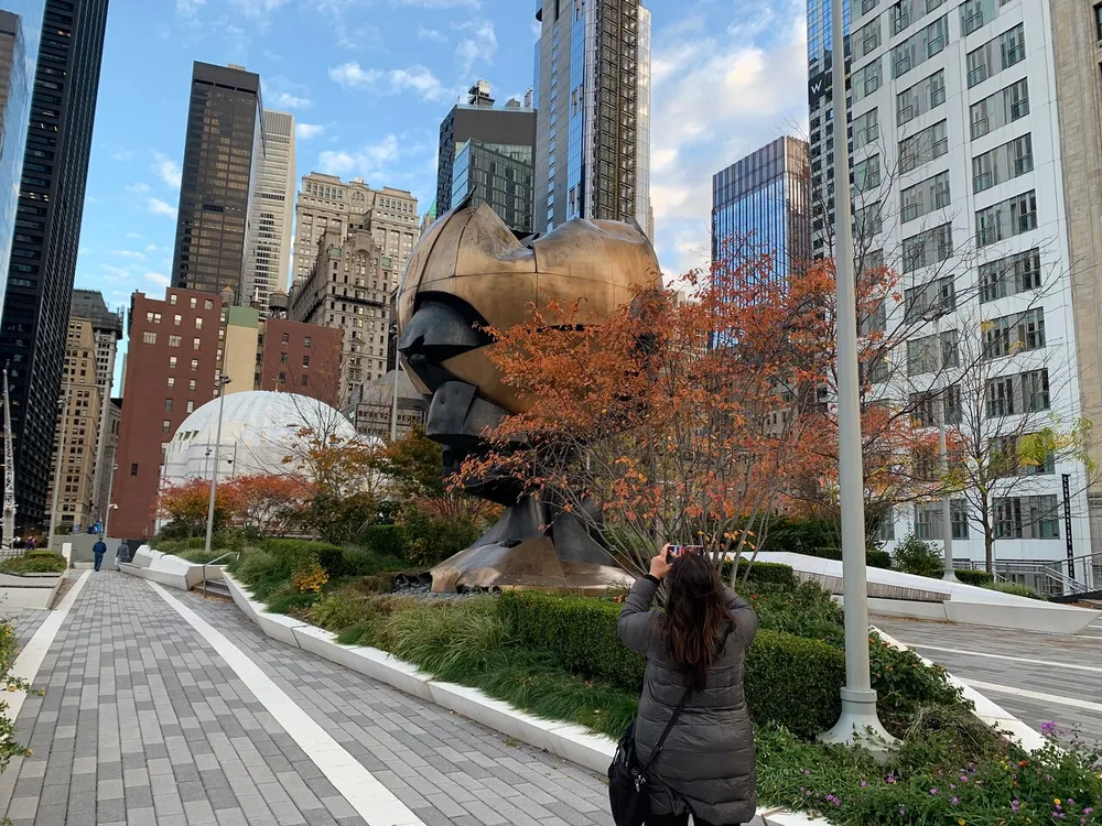 A person is taking a photograph of a large damaged bronze sculpture in an urban park setting with skyscrapers in the background