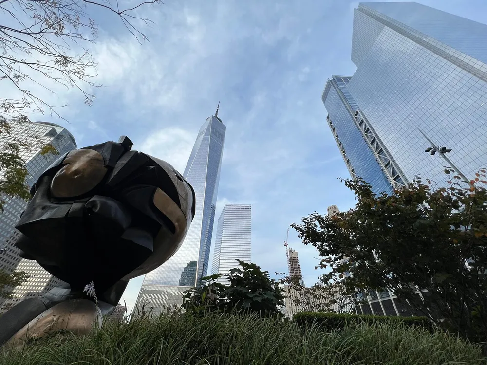 The image shows a low-angle view of a sphere sculpture with a flower attached set against the backdrop of towering skyscrapers and a clear sky