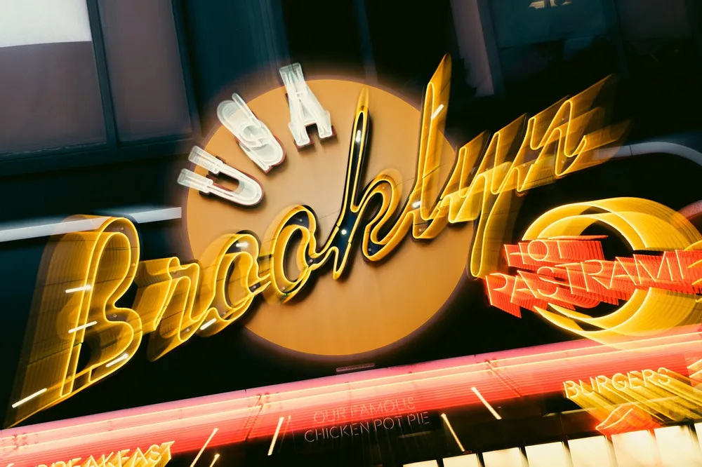 The image shows a neon sign that reads Brooklyn with a motion blur effect and features words like USA Hot Pastrami Burgers and Our Famous Chicken Pot Pie