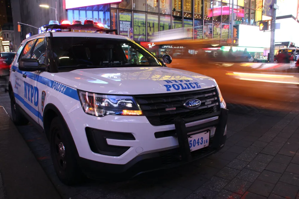 A New York Police Department NYPD vehicle is parked on the street with its lights flashing as the city lights blur in motion at night