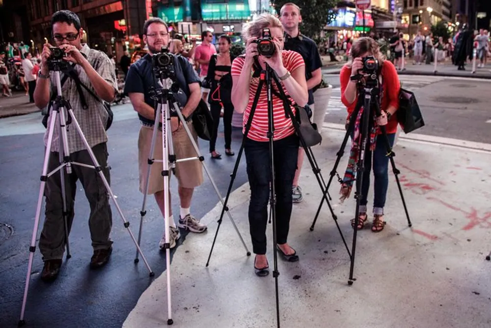 A group of photographers with their cameras mounted on tripods are taking pictures in a bustling urban setting at night
