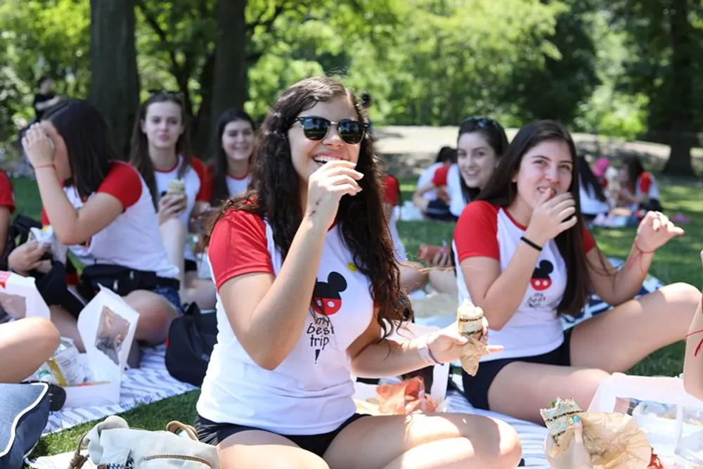 A group of smiling young people wearing matching t-shirts are enjoying a picnic in the park