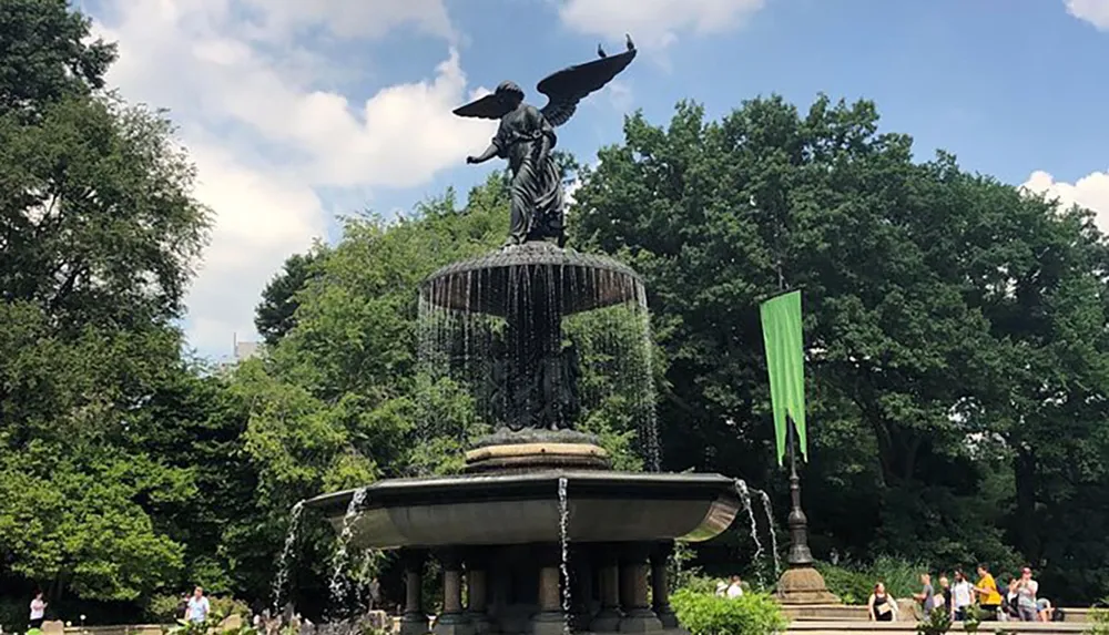 The image depicts a large ornamental fountain featuring a statue of an angel with outstretched wings set against a backdrop of lush trees in a park where people are relaxing on benches