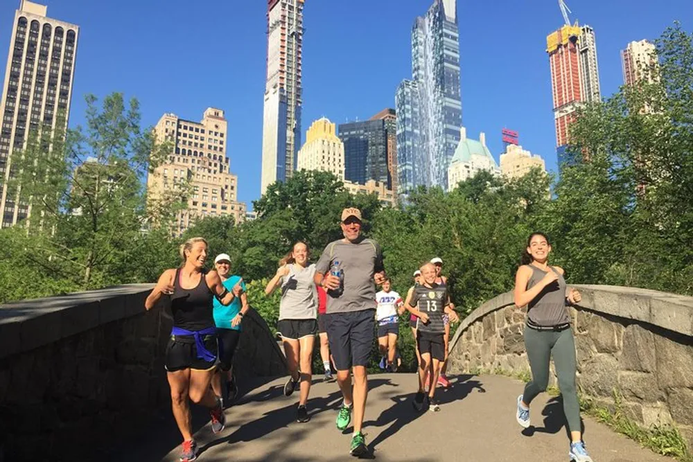 A group of people is jogging together on a path in a park with a backdrop of urban skyscrapers under a clear blue sky