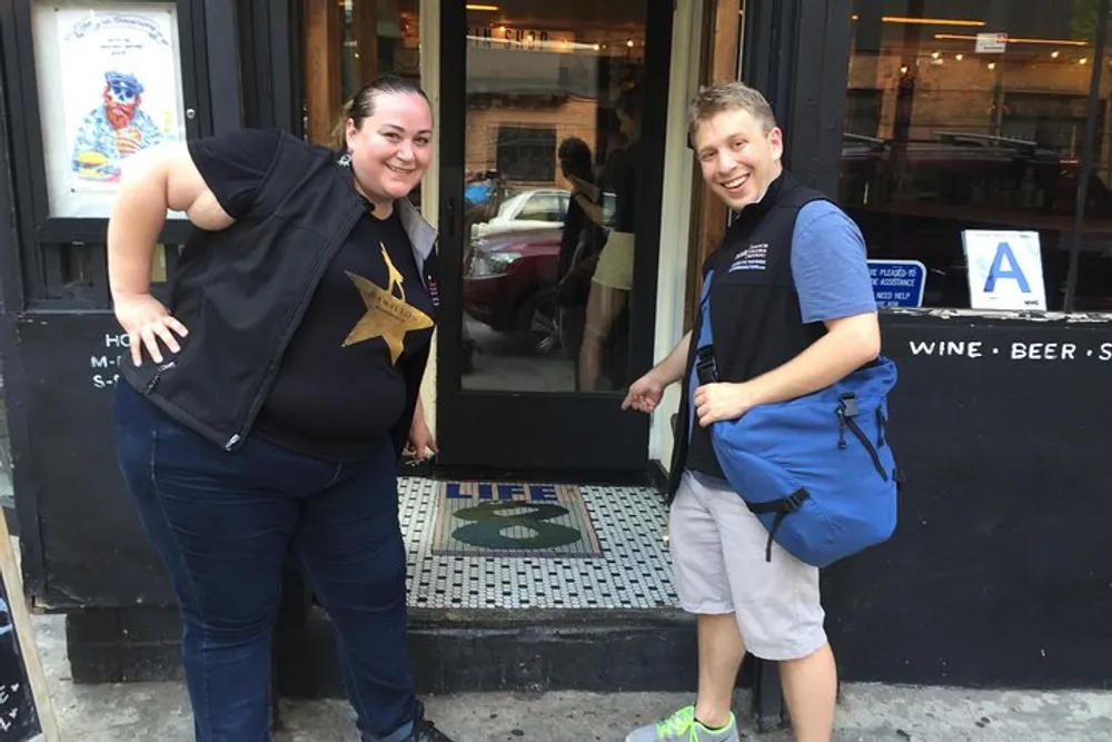 Two individuals are smiling and posing in front of a restaurant door with one person pointing at the entrances floor mosaic