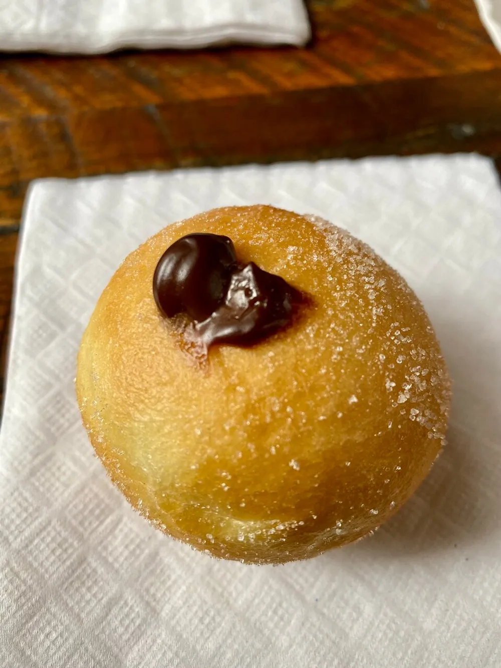 The image shows a single sugary doughnut with a chocolate drop on top resting on a white napkin with a wooden surface in the background