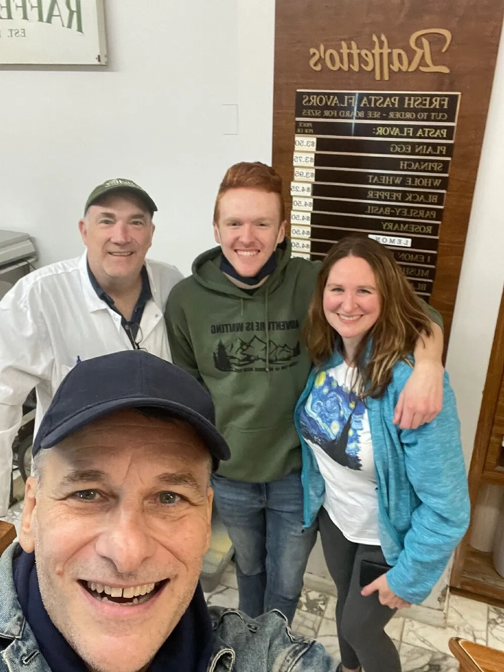 Four people are smiling for a selfie in a room with a wooden board displaying various options in reversed text suggesting theyre at a food service establishment