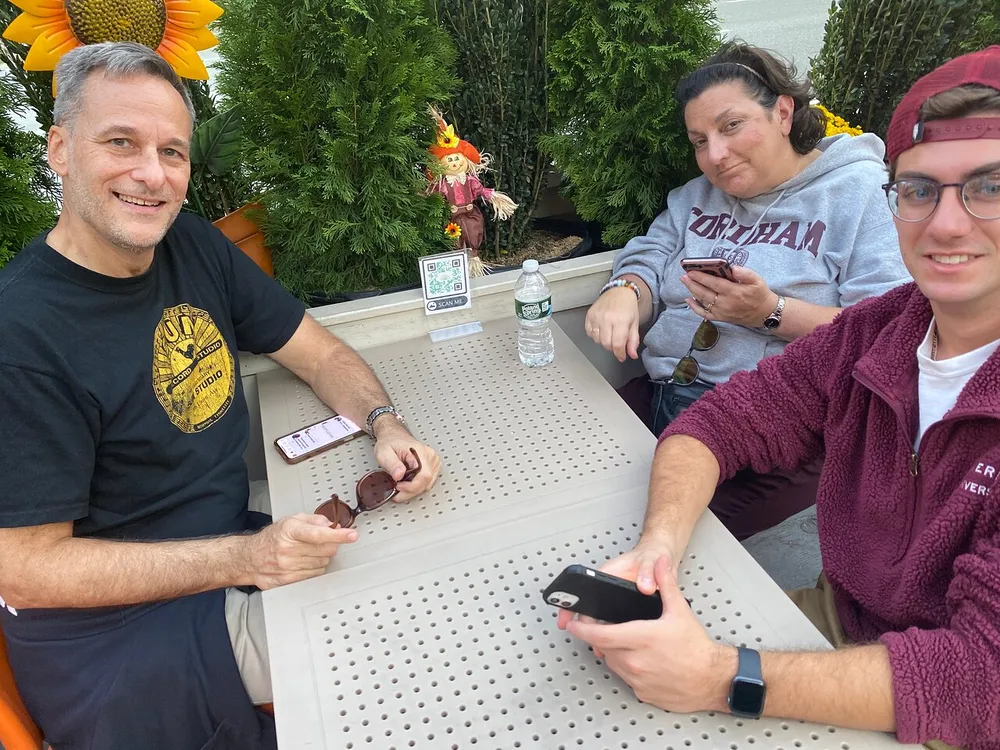 Three people are seated at an outdoor table each holding a smartphone with autumn decorations nearby
