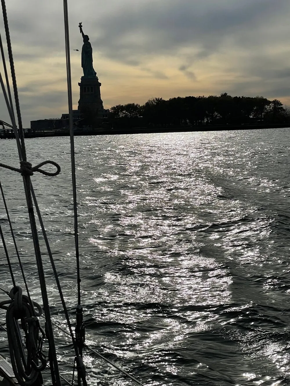 The image captures a view of the Statue of Liberty at sunset from the perspective of a boat with shimmering waters in the foreground and the silhouette of the statue against a cloudy sky in the background