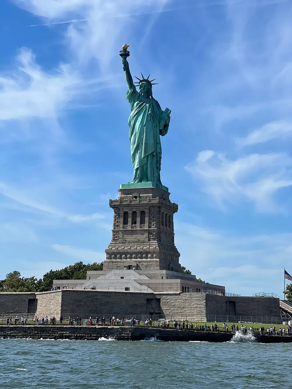 The image captures the Statue of Liberty standing tall against a blue sky with numerous visitors visible at its base